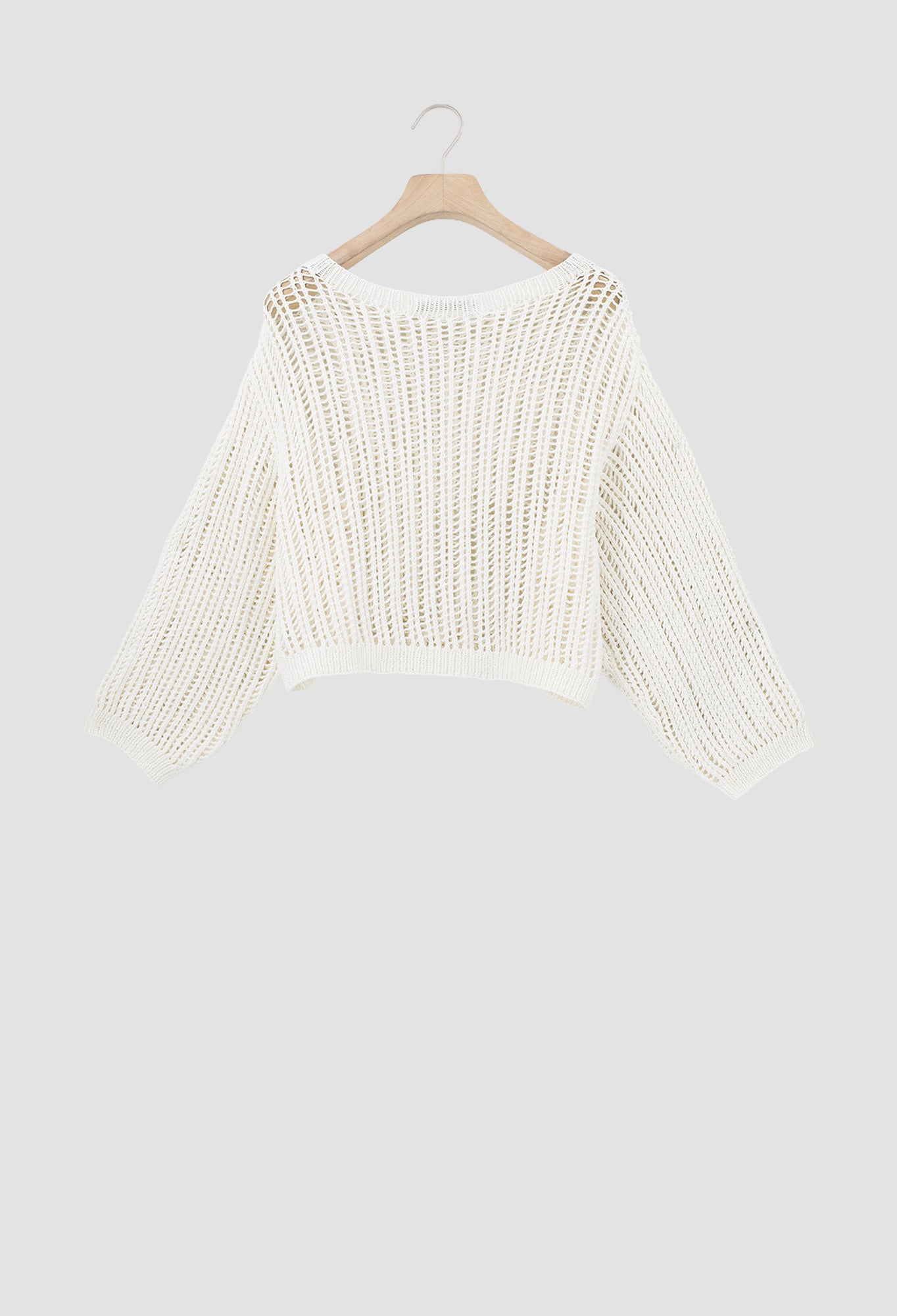 An off-white open-knit cropped sweater with long sleeves, hanging on a wooden hanger against a solid light gray background.