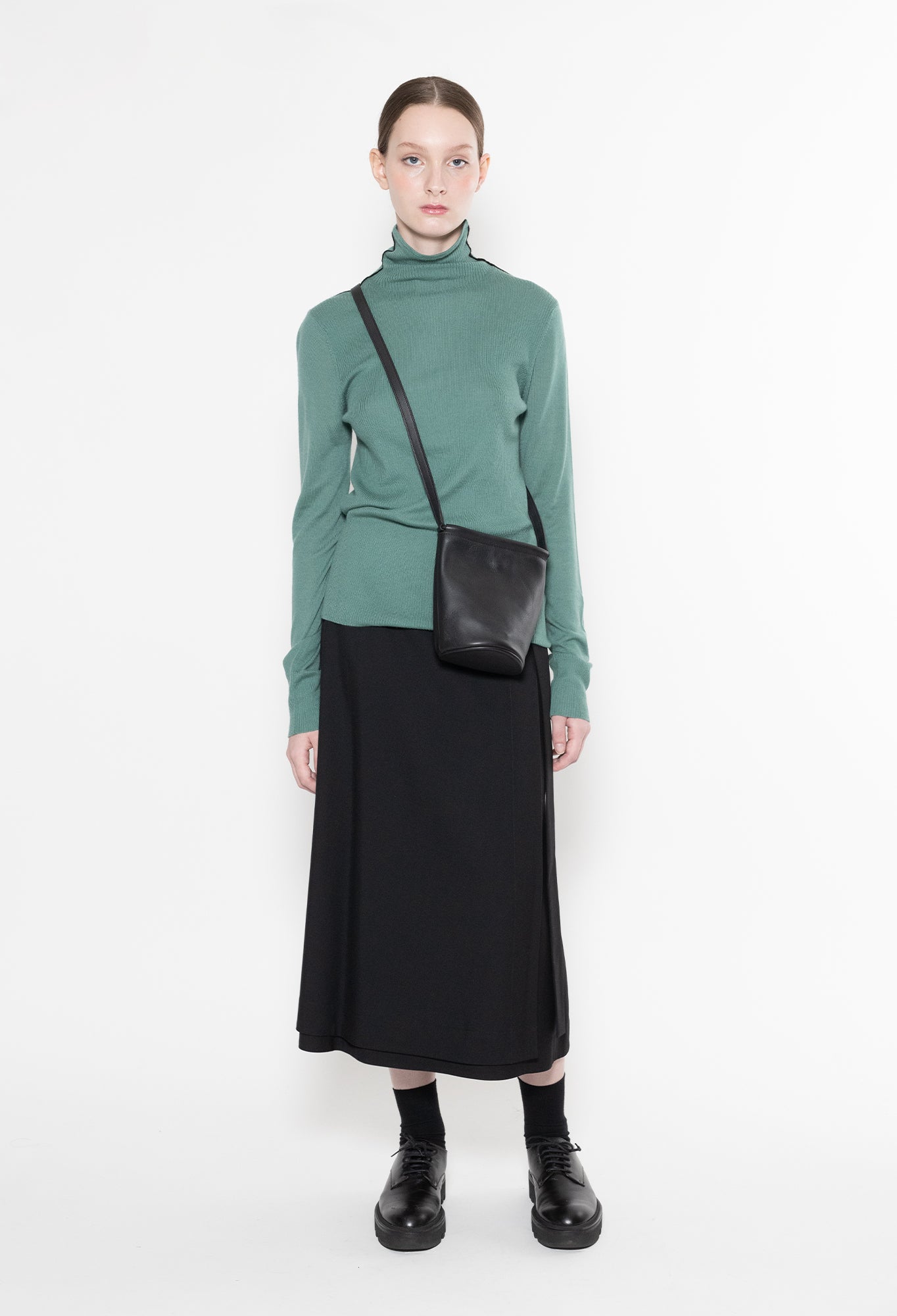 Person wearing a green turtleneck and black skirt with a black bag on a white background
