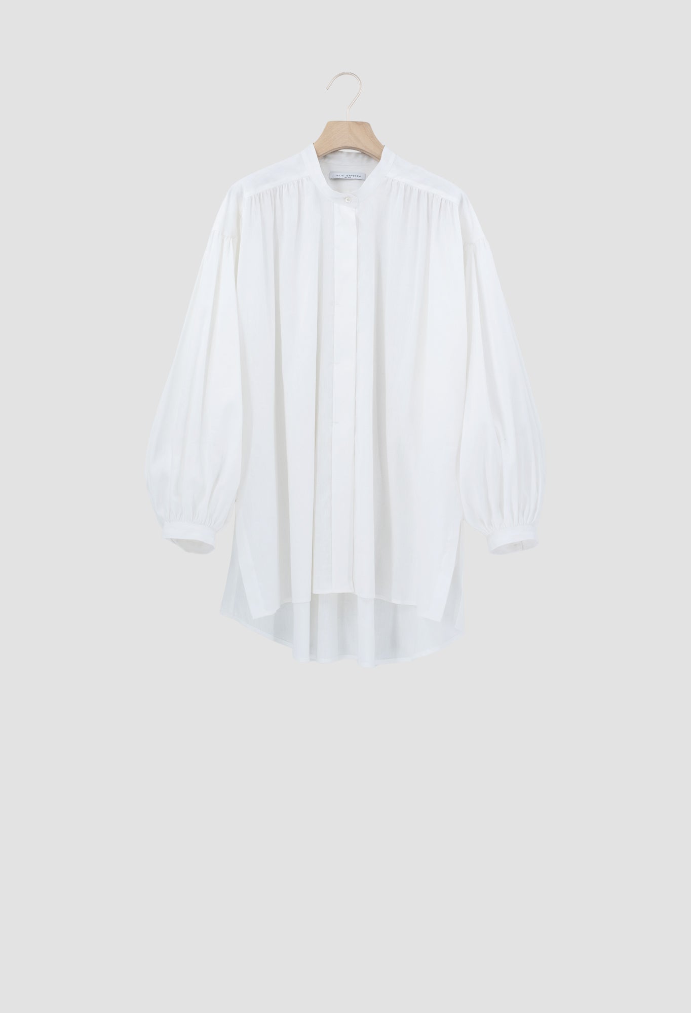 A white long-sleeve blouse with a high-low hem and gathered sleeves, hanging on a wooden hanger against a light gray background.