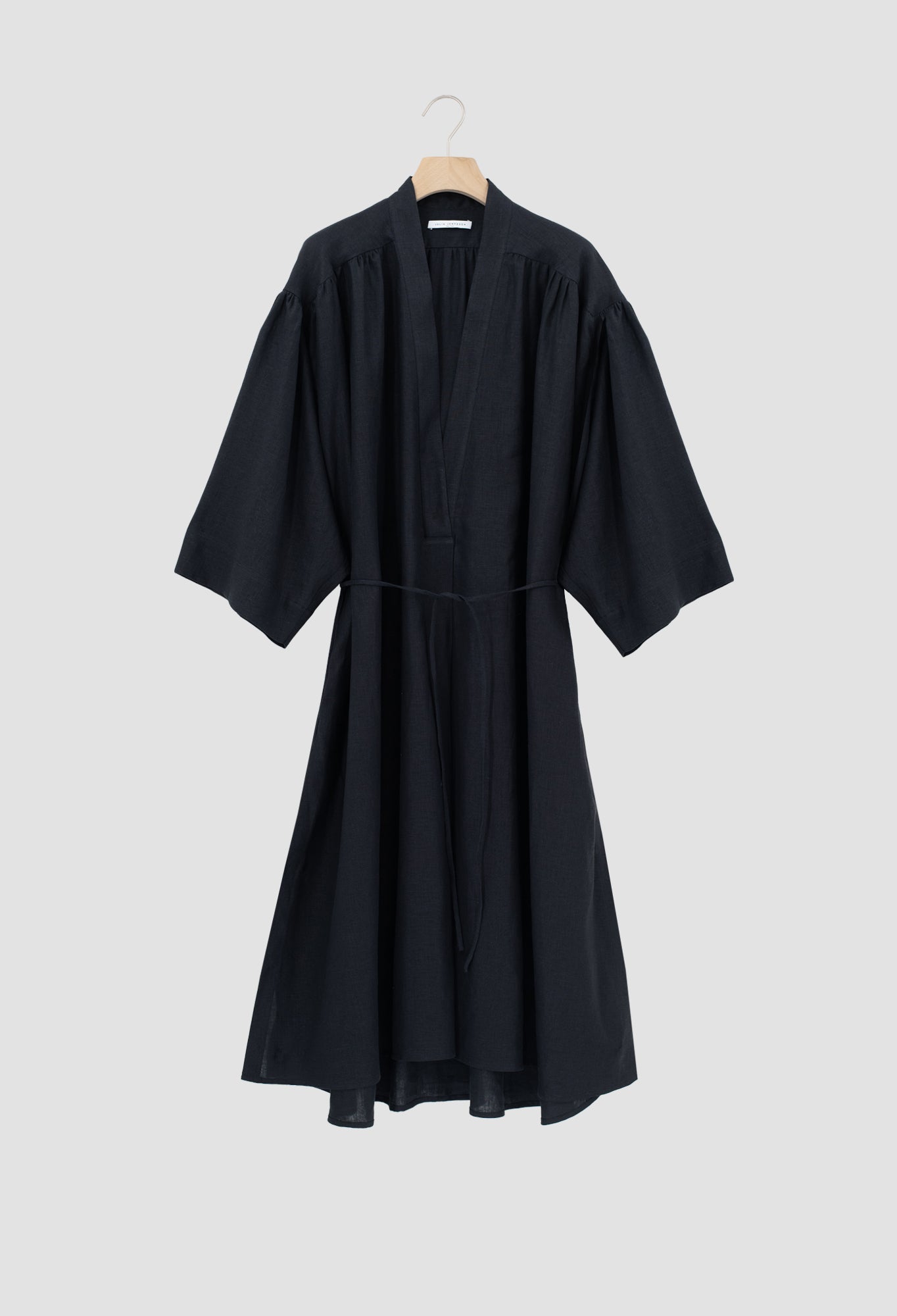 A black wrap-style dress with wide three-quarter sleeves and a self-tie waist belt, hanging on a wooden hanger against a solid light gray background.