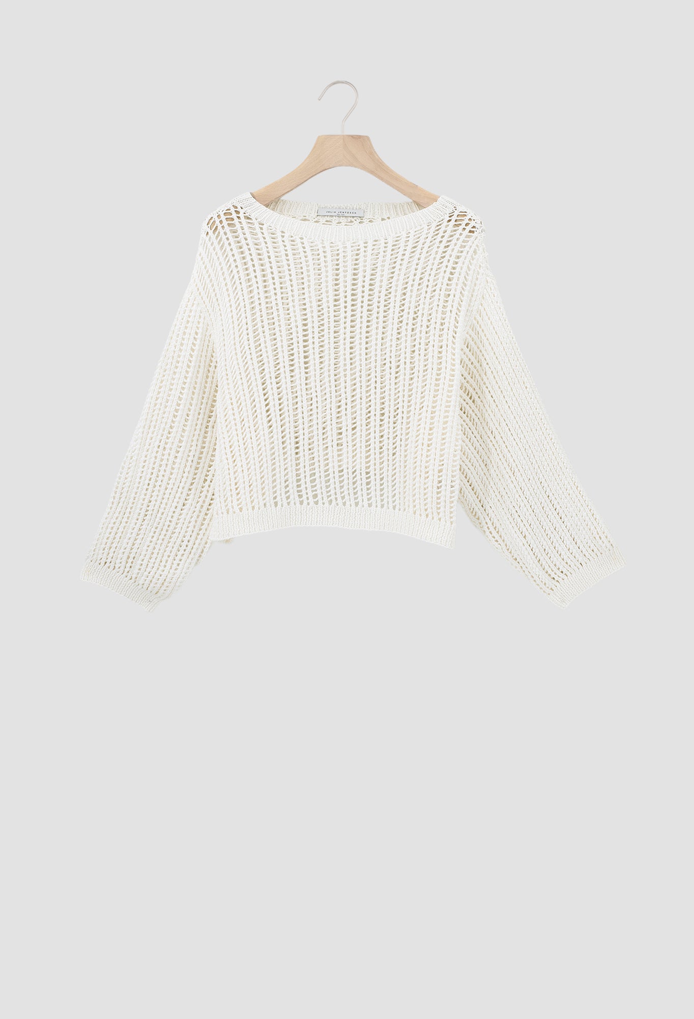 An off-white open-knit cropped sweater with long sleeves, hanging on a wooden hanger against a light gray background.